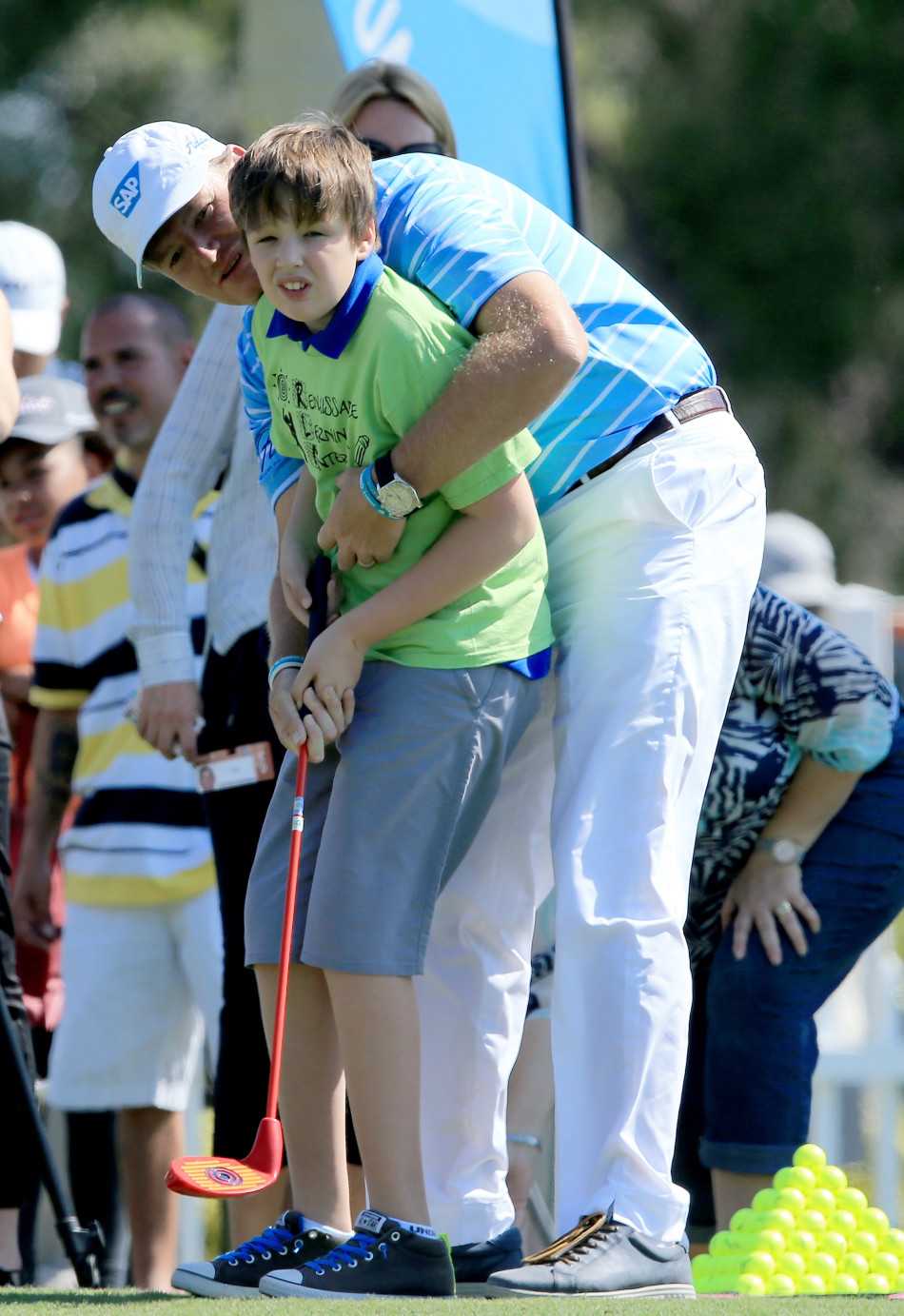 大易哥与查尔默斯夫妇在活动现场开心合影，背景是蓝天绿草和孩子们欢快的身影。