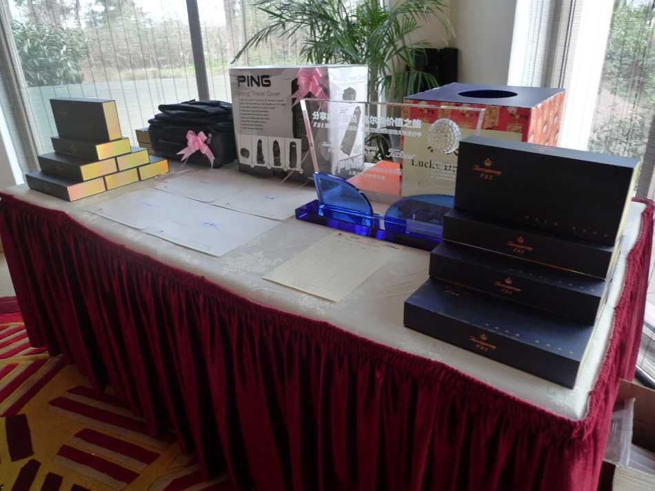 嘉宾微笑着从礼仪手中接过礼盒，背景是开阔的球场，画面充满收获的喜悦与尊贵感