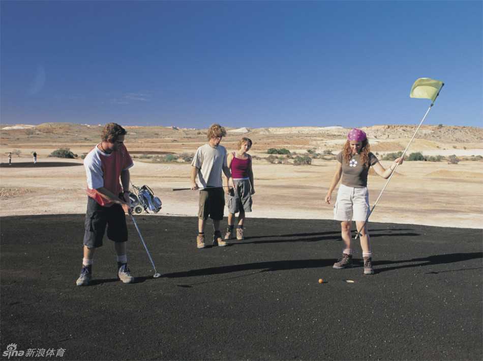 一名高尔夫球手正站在由人工草皮铺设的发球台上，在漫天红沙与嶙峋矿石构成的苍茫背景中准备挥杆。